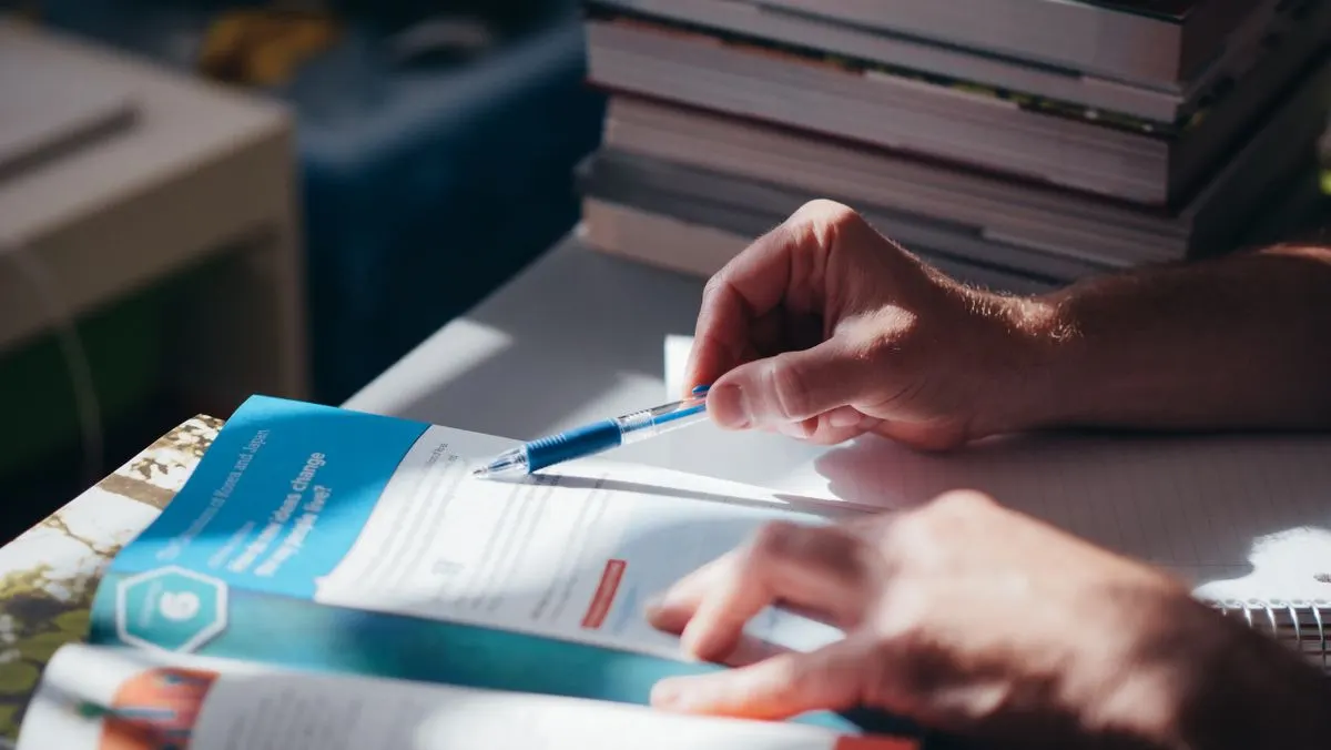 Hands reading and writing on documents at a desk beside books